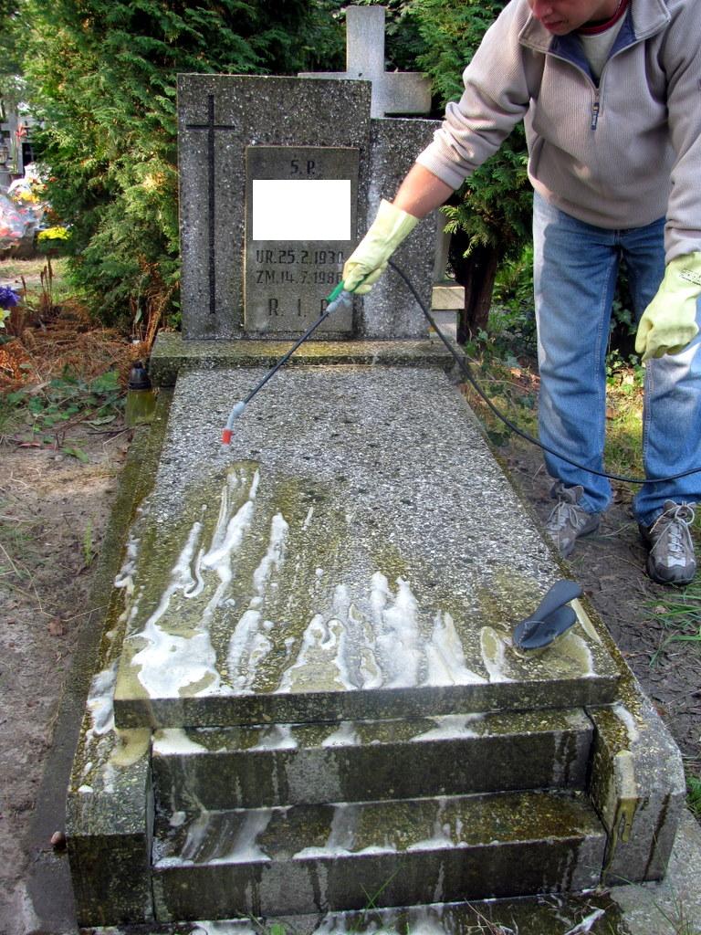 Osoba w rękawicach ochronnych spryskuje nagrobek środkiem czyszczącym, widoczne białe smugi preparatu na kamiennej powierzchni.